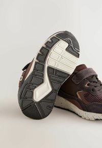 Brune sportsko med en struktureret overdel, en velcro-rem og en hvidprikket mellemsål. Gummisålen har et mønster for godt greb.