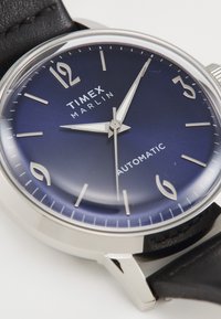 Ceasul Timex Marlin are un carcasă din oțel inoxidabil argintiu, un cadran albastru închis cu marcaje orare argintii și o curea din piele neagră.