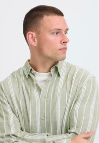 Chemise boutonnée à rayures vert clair avec des lignes blanches, col classique, coupe décontractée et texture légère, portée par-dessus un t-shirt blanc à col rond.