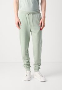Pantalones de chándal verde claro con cinturilla elástica y cordón, que cuentan con bolsillos laterales y una pequeña etiqueta roja en la parte delantera.