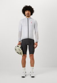 Veste de cyclisme légère blanche avec fermeture éclair intégrale, shorts noirs et casque gris. Texture lisse, coupe athlétique et détails de logo visibles.