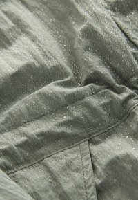 Water-resistant fabric in muted green, featuring textured surface with droplets, visible stitching, and smooth texture indicating flexibility.