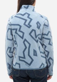 Eine Person trägt eine hellblaue Fleecejacke mit dunkelblauen abstrakten geometrischen Mustern, kombiniert mit einer dunklen Jeans, von hinten gesehen.