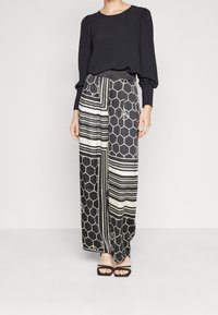 Haut noir à manches longues avec épaules bouffantes, associé à un pantalon à jambes larges présentant un motif géométrique noir et blanc. Sandales à talons noires.