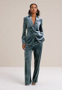 Sammetssuit i dämpad grön, med en figursydd kavaj med spetsiga slag och vida byxor. Slät textur, minimal detaljering.