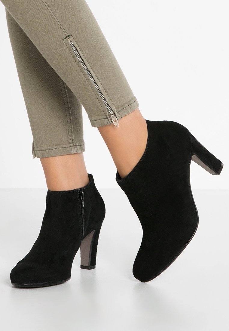 Bottes à talons hauts en daim noir portées avec un pantalon beige doté de fermetures éclair argentées sur les côtés.