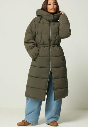 Femme portant un long manteau matelassé vert olive avec un col montant, un jean bleu à jambes larges et des chaussures marron, debout devant un fond uni.