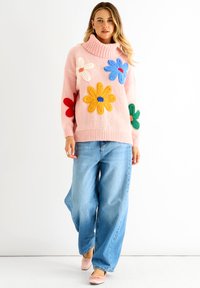 Pink knitted sweater with multicolored floral appliqués, ribbed high neck, paired with loose blue jeans and light pink ballet flats.