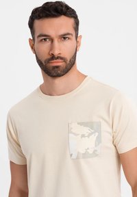 Camiseta de algodón beig con cuello redondo y un bolsillo a parches estampado que presenta un diseño de camuflaje claro en tonos de blanco y gris.
