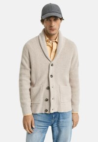 Man som bär en grå keps, beige sjalkrage stickad cardigan med knappar och fickor, ljusorange skjorta och blå jeans, står mot en vit bakgrund.