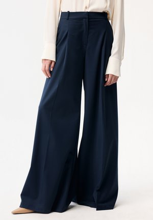 Kobieta ubrana w granatowe spodnie z wysokim stanem i szerokimi nogawkami, zestawione z kremową bluzką z długim rękawem oraz beżowymi szpiczastymi butami.