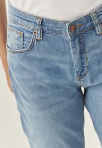 Jeans din denim albastru deschis cu o croială slim, având accesorii din metal cu un finisaj cupru, cusături portocalii și un design cu cinci buzunare. Textură netedă.