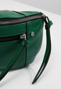 Sac en cuir vert texturé avec fermeture éclair argentée et bouton magnétique, doté de deux languettes en cuir pour fermeture éclair et d'une sangle de poignet assortie.