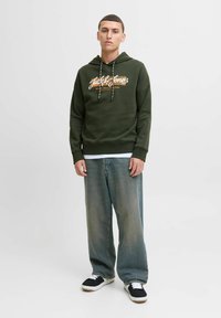 Grøn hættejakke med et hvidt og orange logo, snore i hætten og ribbestrikket kant, parret med lyseblå baggy jeans og sorte sneakers.