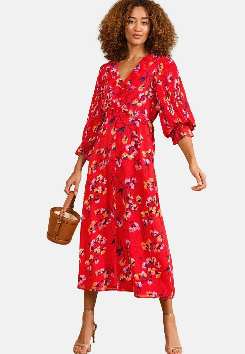 Rochie florală roșie cu decolteu în V, detalii ondulate și mâneci bufante trei sferturi. Prezintă flori galbene și mov pe un material fluid.
