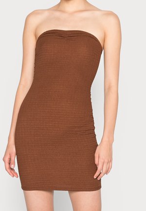 Femme portant une mini-robe marron texturée, sans bretelles et moulante, debout devant un fond clair uni.