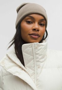 Kreemi värvi puffjakki, millel on teemantkujuline tikand ja kõrge krae, millel on mansetid ja beež villane beanie.