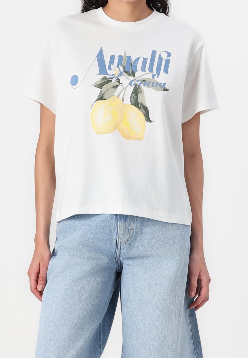Camiseta blanca de algodón con texto azul "Amalfi Coast" y gráfico de limón amarillo, con un corte relajado y mangas cortas.