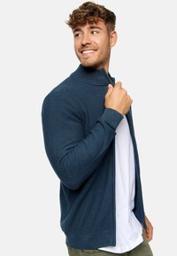 Cárdigan texturizado azul marino con cuello alto, cierre de cremallera y mangas largas, superpuesto a una camiseta blanca. Contiene detalles de canalé.