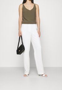 Top sin mangas de color verde oliva con tirantes decorativos, pantalones anchos blancos y un bolso negro. Sandalias de tacón blancas completan el look.
