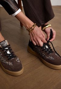 Brown suede sneakers with leopard print accents, black laces, and gold hardware. Styled with brown pants and a leather jacket.