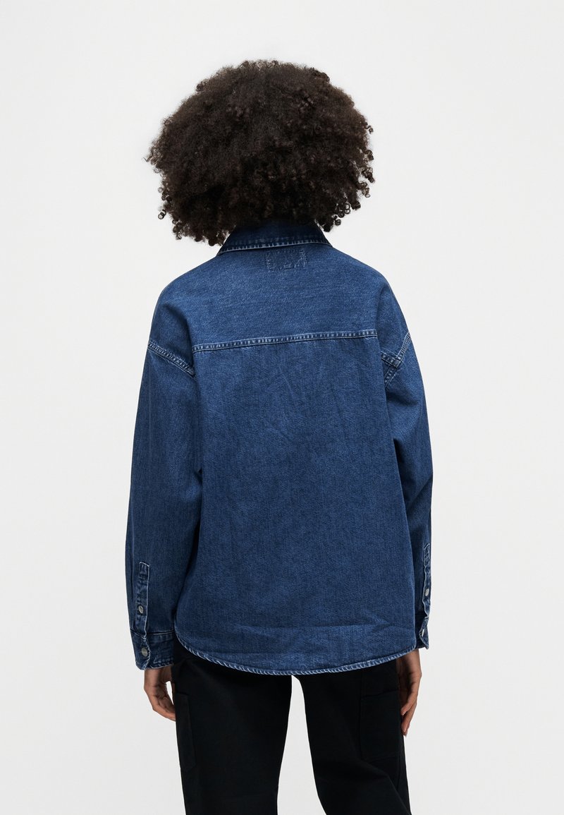 Denim jacket in dark blue, with long sleeves and a buttoned cuff. Features a classic collar and back yoke detailing, showing textured fabric.