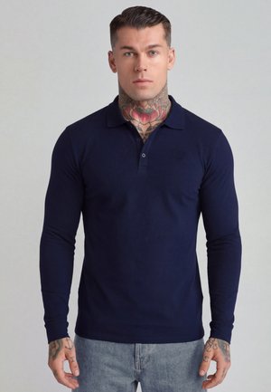 Mand med tatoveringer på nakke og hænder iført navyblå langærmet polo og lyseblå jeans, set forfra mod en ensfarvet baggrund.