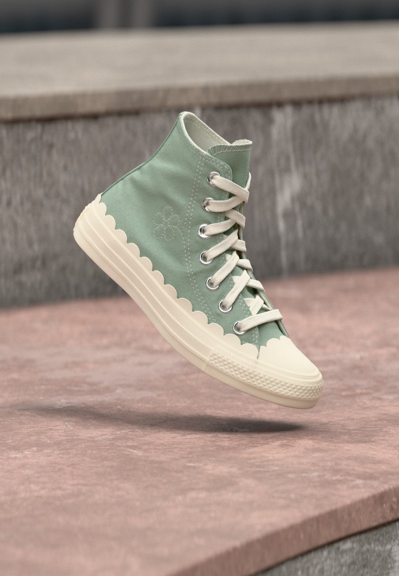 Πράσινο καμβαδένιο ψηλό παπούτσι με λευκά κορδόνια και κρεμ σόλα με κυματιστό σχέδιο, εμφανίζεται στον αέρα πάνω από σκαλοπάτια από πέτρα.
