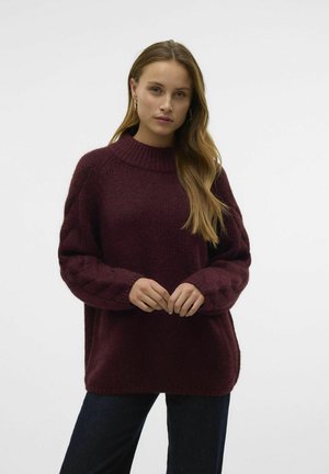 Viininpunainen ylipitkä neulepaita korkealla kaula-aukolla ja päälikudontaa hihoissa. Pehmeä, texturoitu kangas yhdistettynä tummaan farkkuun.