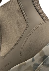 Stivaletto impermeabile con parte superiore in tela beige, dettagli testurizzati e suola patterned. Presenta cuciture visibili e un bordo in gomma liscia.
