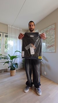 Svart t-shirt förpackad i genomskinlig plast med etiketter fästa. Modell som håller t-shirten står i ett välinredd rum med inomhusväxter.