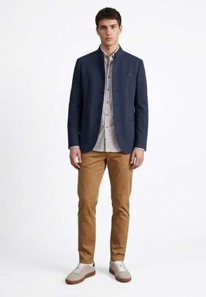 Joven de pie, vistiendo un blazer azul marino, camisa de botones con estampado, pantalones color beige y zapatillas grises claras frente a un fondo blanco liso.