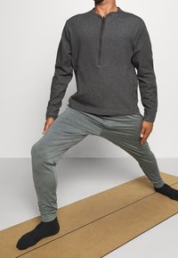 Sweat-shirt gris à col zippé et joggers gris clair, avec une coupe décontractée. Portés lors d'une pose de yoga sur un tapis beige. Chaussettes noires visibles.