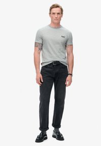T-shirt grigio a maniche corte con logo, abbinato a jeans neri a gamba dritta. Gli accessori includono una cintura nera e scarpe nere con dettagli.