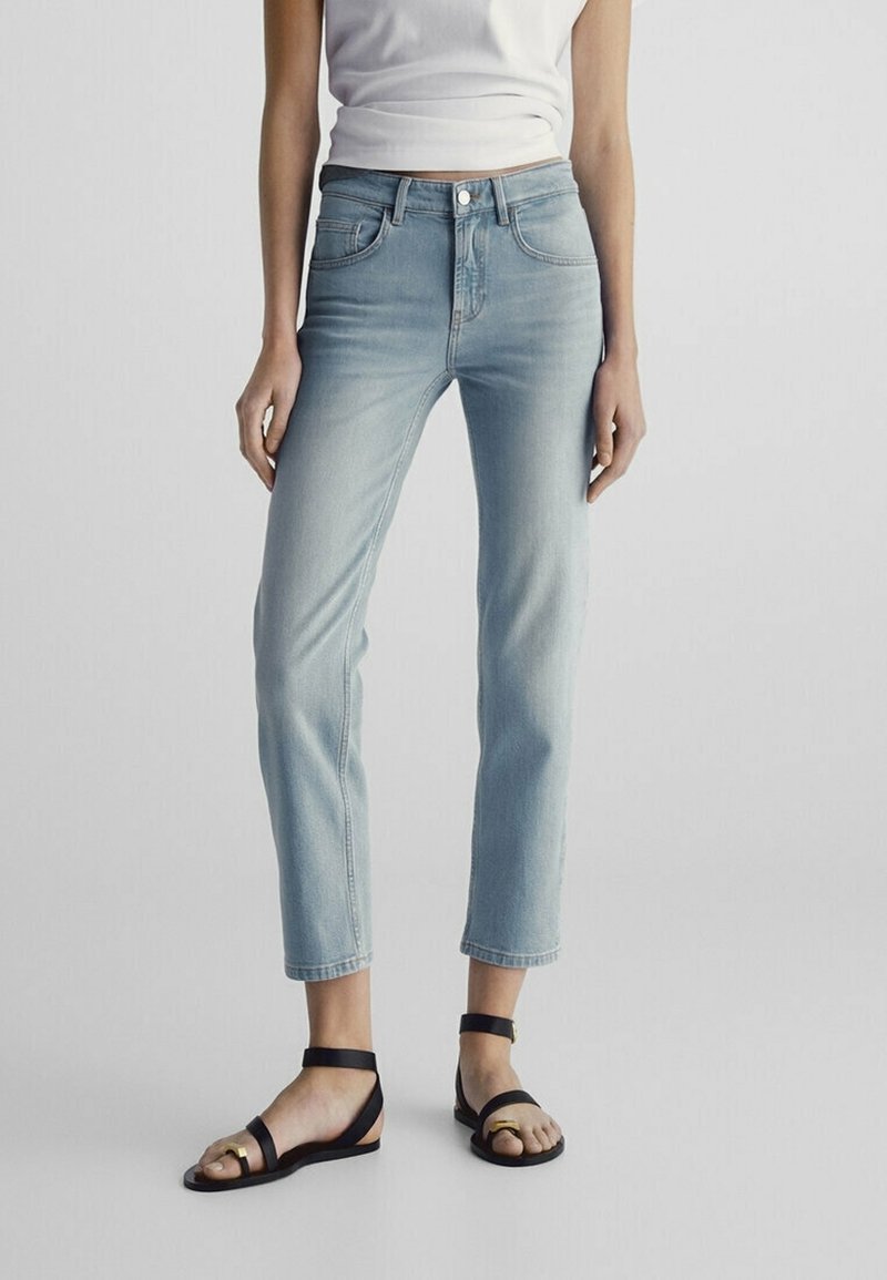 Light blue cropped jeans in denim, five-pocket design, high-rise waist, paired with black flat sandals featuring a gold accent on the strap.