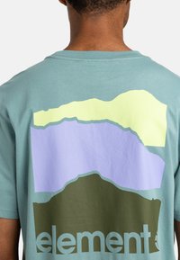 Lys teal t-shirt med et stort, flerfarvet abstrakt grafik på ryggen med lag af grøn, lilla og gul.