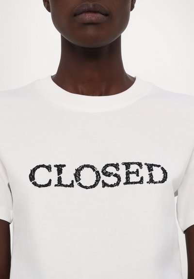 Balta kokvilnas T-kreklā ar melnu izšūtu tekstu "CLOSED" uz krūšu, klasiskā apaļā kakla izgriezuma un īsām piedurknēm.