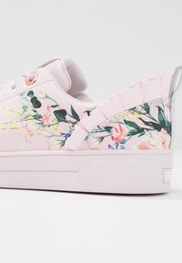 Baskets en tissu rose avec motif floral, semelle texturée, volants décoratifs au niveau du talon et œillets en or rose. Finition lisse et design à profil bas.