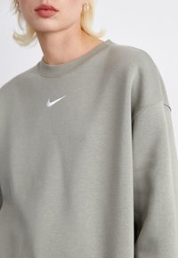 Sweatshirt cinza claro feito de tecido macio, apresentando um decote canelado e um pequeno logotipo branco da Nike no peito. Corte descontraído, design minimalista.