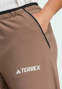 Brązowe sportowe spodnie z elastycznym pasem, czarnymi akcentami, bocznymi kieszeniami i białym logo "TERREX" na dolnej części lewej nogawki. Gładka tekstura.