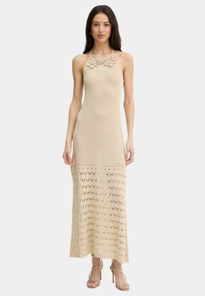 Donna che indossa un abito maxi beige senza maniche in crochet con dettagli floreali sulla scollatura e motivo a pizzo aperto sulla metà inferiore.