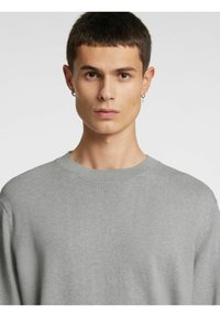 Lys grå strikkegenser med rund hals og ribbestrikkede mansjetter. Glatt tekstur, avslappet passform og minimale sømmer synlige på skulderen.