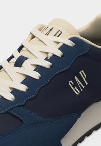 Ναυτικό μπλε και κρεμ αθλητικό παπούτσι με λευκά κορδόνια, υφασμάτινα μπλε καλύμματα και λογότυπο "GAP" στο πλάι και στη γλώσσα.