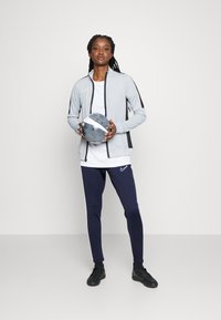 Giacca sportiva grigia con accenti neri, maglietta bianca, pantaloni blu navy con righe bianche e scarpe da ginnastica nere, mentre tiene in mano un pallone da calcio grigio.