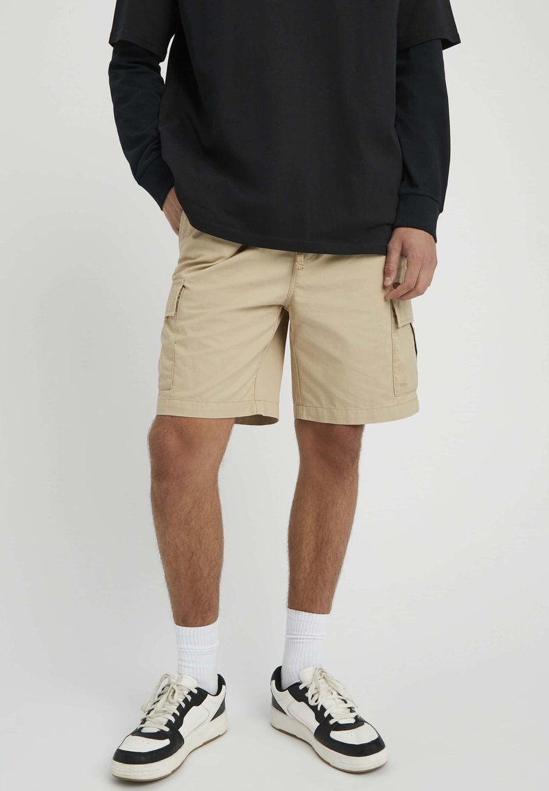Shorts cargo couleur kaki avec deux poches latérales et une coupe droite, assortis à un t-shirt noir oversized et des baskets blanches de sport.