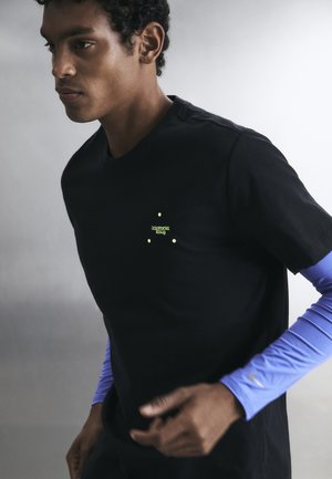 Νέος άνδρας φοράει μαύρο μπλουζάκι πάνω από ένα μακρυμάνικο ανοιχτό γαλάζιο πουκάμισο, ποζάροντας με συγκεντρωμένη έκφραση σε απλό φόντο.