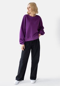 Fioletowy, przytulny sweatshirt z opuszczonymi ramionami, w luźnym kroju, z prążkowanymi mankietami, zestawiony z czarnymi spodniami typu wide-leg i białymi sneakersami.