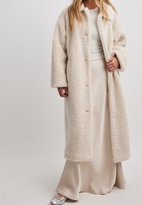 Beige kunstpelsjakke med krave, lange ærmer og en afslappet pasform. Har knaplukning og blød, fluffy tekstur. Sammen med en cremefarvet nederdel.