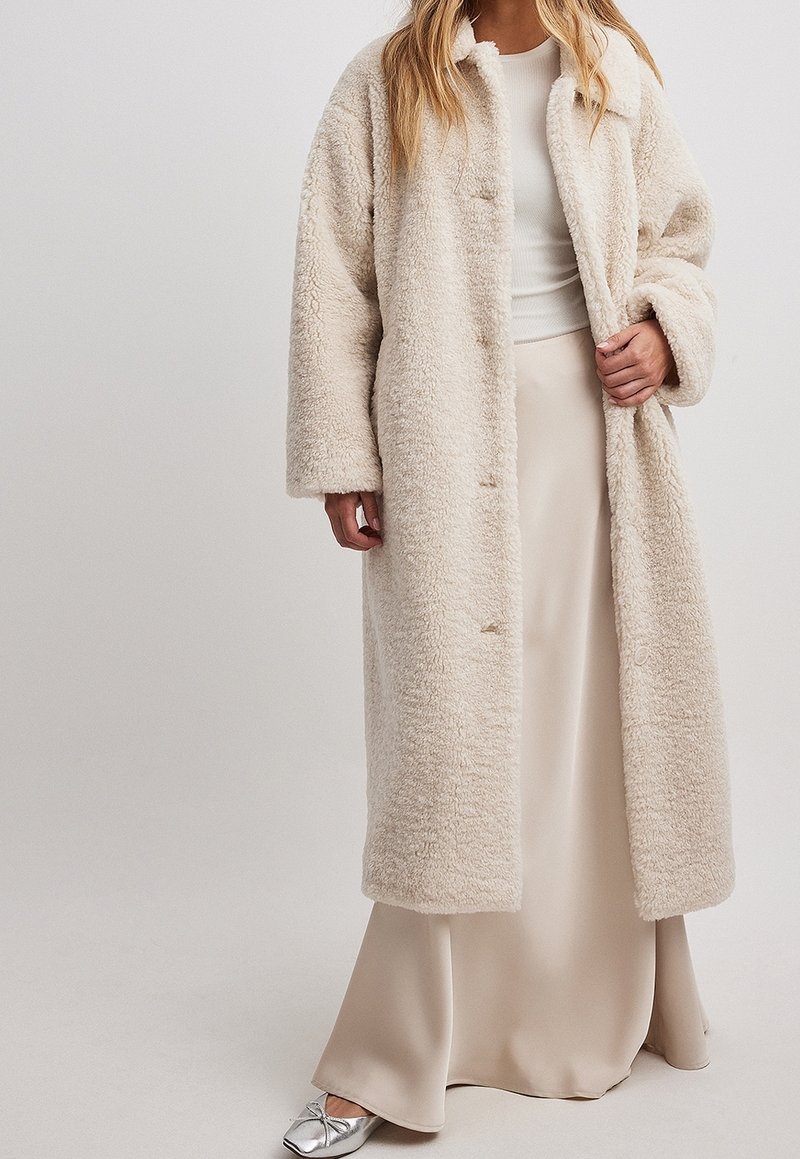 Beige kunstpelsjakke med krave, lange ærmer og en afslappet pasform. Har knaplukning og blød, fluffy tekstur. Sammen med en cremefarvet nederdel.