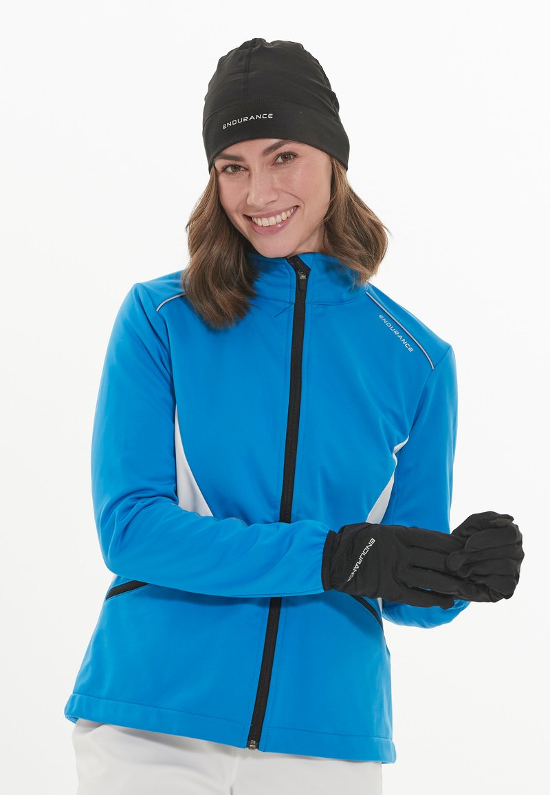 Femme en veste zippée bleue et chapeau noir mettant des gants noirs, souriante, sur fond blanc